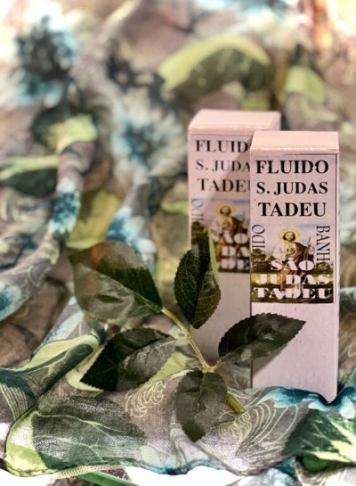 Duas embalagens retangulares de fluido São Judas Tadeu com folhas verdes falsas e tecido estampado ao fundo.