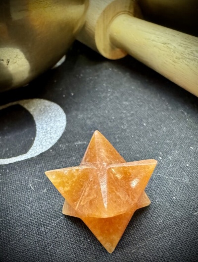 Cristal laranja em forma Merkaba sobre tecido preto com padrão branco ao lado de objeto metálico e bastão de madeira
