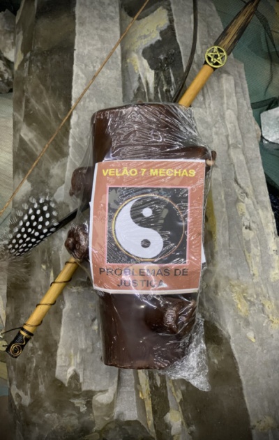 Vela castanha com etiqueta yin-yang embalada em plástico sobre pedras, com pena e varinha decorativa ao lado.