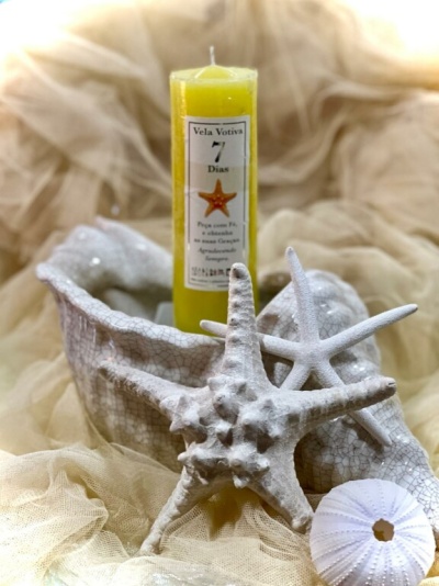 Vela votiva amarela com rótulo branco dentro de um objeto decorativo branco com estrelas-do-mar brancas ao redor, sobre tecido bege.