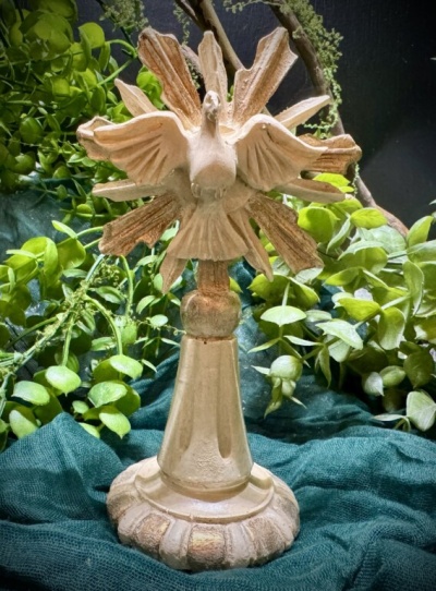 Escultura dourada de pomba com asas abertas sobre base redonda, decorada com folhagem verde ao fundo