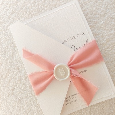 Convite branco com laço cor-de-rosa e selo branco sobre tecido claro