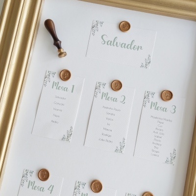 Quadro organizado com cartões de design branco, verde e preto, selados com cera dourada para números e nomes de mesas.