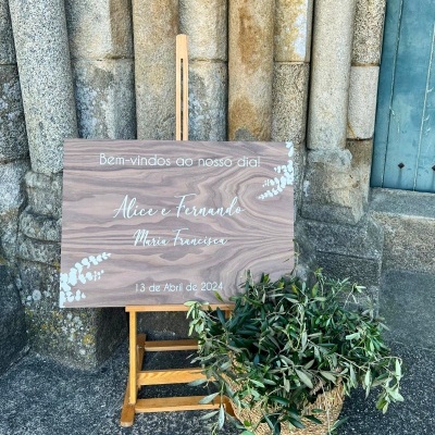 Placa decorativa de madeira com texto de boas-vindas para evento, apoiada em cavalete junto a vaso de plantas, fundo de parede de pedra.