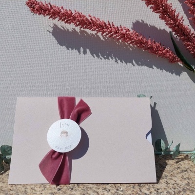 Envelope bege com fita vermelha e selo branco sobre superfície de pedra com flores secas ao redor.