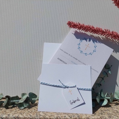 Convite branco com etiqueta e cordão azul, folhas verdes e ramo vermelho seco sobre superfície bege.