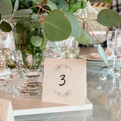 Cartão de mesa número 3 com padrão floral e jarro de vidro com folhas verdes sobre mesa com taças