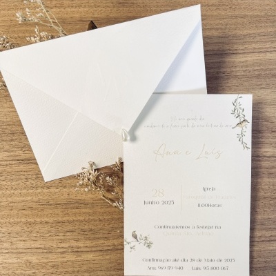 Convite de casamento branco com envelope branco e detalhes florais sobre madeira.