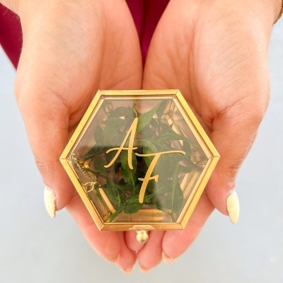 Caixa hexagonal de vidro dourada com ramos verdes e letras douradas AF, nas mãos.
