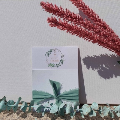 Convite branco com laço verde e etiqueta decorativa, folhas de eucalipto e planta vermelha seca