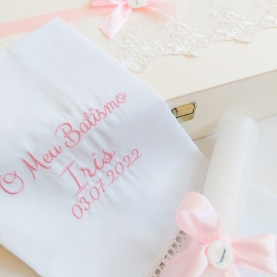 Conjunto de batismo com toalha branca bordada, vela decorada e caixa com renda e laço cor de rosa.