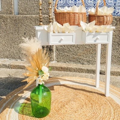 Mesa branca com cestas de vime, castiçais dourados, garrafa verde com flores secas e tapete redondo de palha