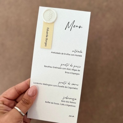 Menu em papel branco com texto em preto e selo branco, segurado por mão com unhas nude