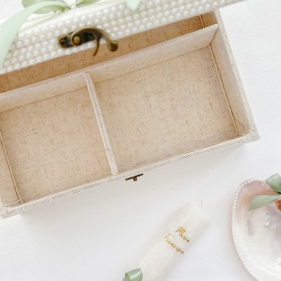 Caixa aberta de tecido bege, vela branca decorada e prato branco com flor e fita verde numa superfície branca
