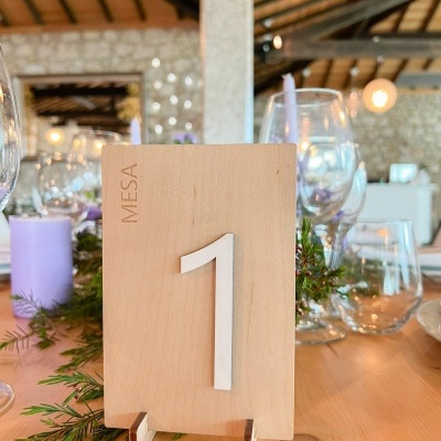 Número de mesa em madeira clara com copos de vidro e decoração verde numa mesa rústica