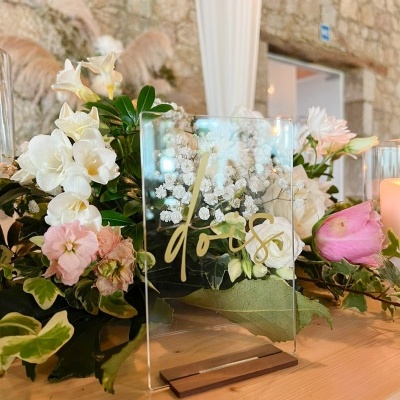 Placa transparente com palavra 'dois' em dourado numa base de madeira, rodeada por flores brancas e rosas sobre mesa de madeira