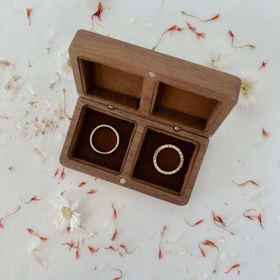 Caixa de madeira aberta com dois anéis dourados interiores sobre superfície branca com flores secas