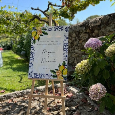 Placa de boas-vindas ao casamento com moldura de azulejos e limões num cavalete de madeira num jardim com flores.