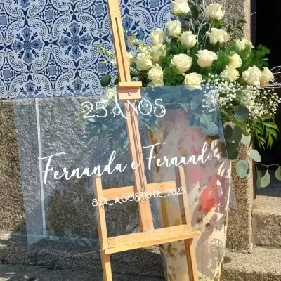 Suporte de madeira com painel transparente escrito e arranjo de flores brancas ao lado, azulejos azuis e parede de pedra ao fundo