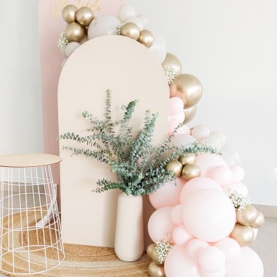 Composição decorativa com painel neutro, balões rosas e dourados, vaso branco com folhagem e banco com estrutura metálica