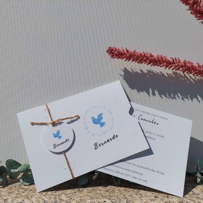 Convite branco com laço de cordão rústico e ilustração azul de pomba, acompanhado de cartão de agradecimento, sobre superfície de pedra com vegetação.