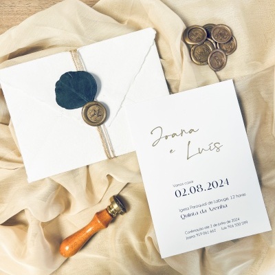 Convite de casamento branco com selo de cera castanha e folha verde sobre pano bege e fundo de madeira clara