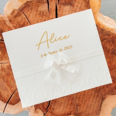 Convite branco com laço e texto dourado colocado sobre uma superfície de madeira.