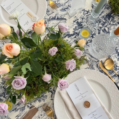 Mesa posta com pratos brancos, talheres dourados, copo de vidro, velas, guardanapo com menu, flores e toalha estampada