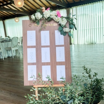 Painel de madeira decorado com flores e folhas verdes, com papéis brancos organizados, em interior com chão de madeira e cortinas brancas.