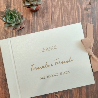 Livro branco com gravação dourada e laço bege ao lado em mesa de madeira com suculentas