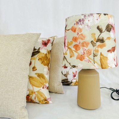 Candeeiro de mesa castanho claro com abajur floral e almofadas decorativas coloridas em fundo branco.