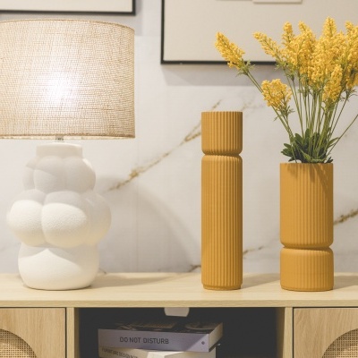 Abajur branco com cúpula de tecido e dois vasos castanhos com flores amarelas num móvel de madeira clara