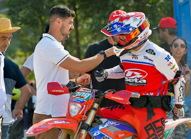 Motociclista em fato Beta vermelho, branco e azul junto a mota de motocross vermelha e pessoas ao redor