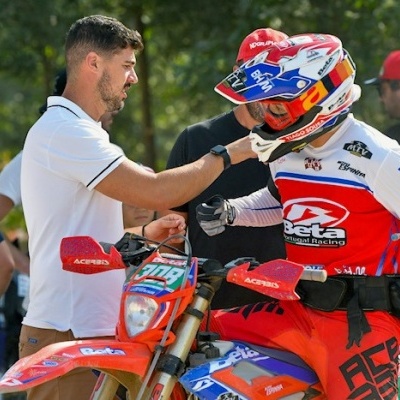 Motociclista em fato Beta vermelho, branco e azul junto a mota de motocross vermelha e pessoas ao redor
