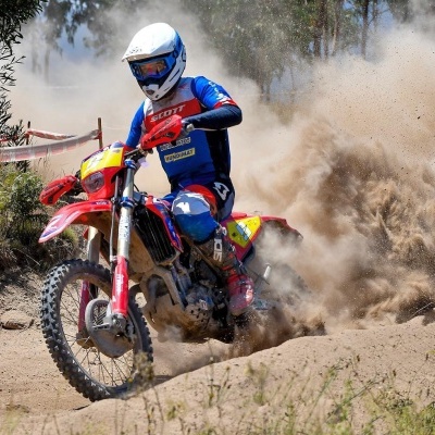 Motociclista com capacete e equipamento azul e vermelho numa moto vermelha a levantar poeira na pista de terra