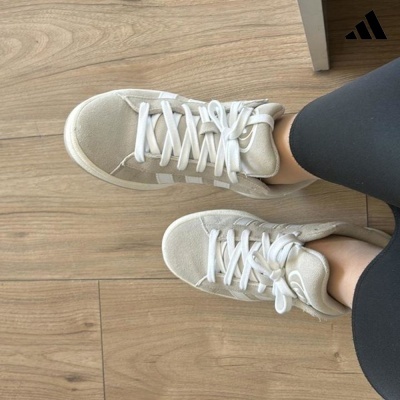 Ténis Adidas camurça bege claro com atacadores brancos, sola branca e três riscas laterais brancas, calças pretas, chão madeira clara