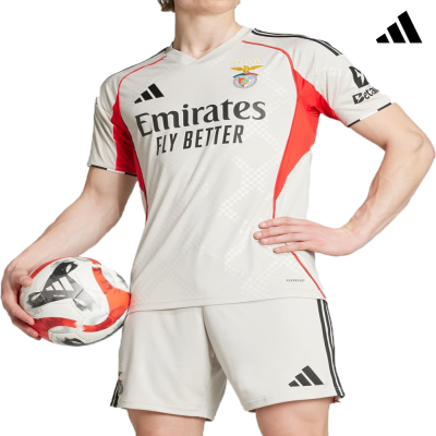 Conjunto de futebol bege com detalhes vermelhos e pretos, logótipos Benfica, Emirates e Adidas, com bola de futebol Adidas nas mãos.