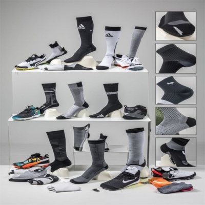 Meias desportivas em preto, branco e cinzento com sapatilhas Adidas, Nike e Mizuno num cenário neutro.