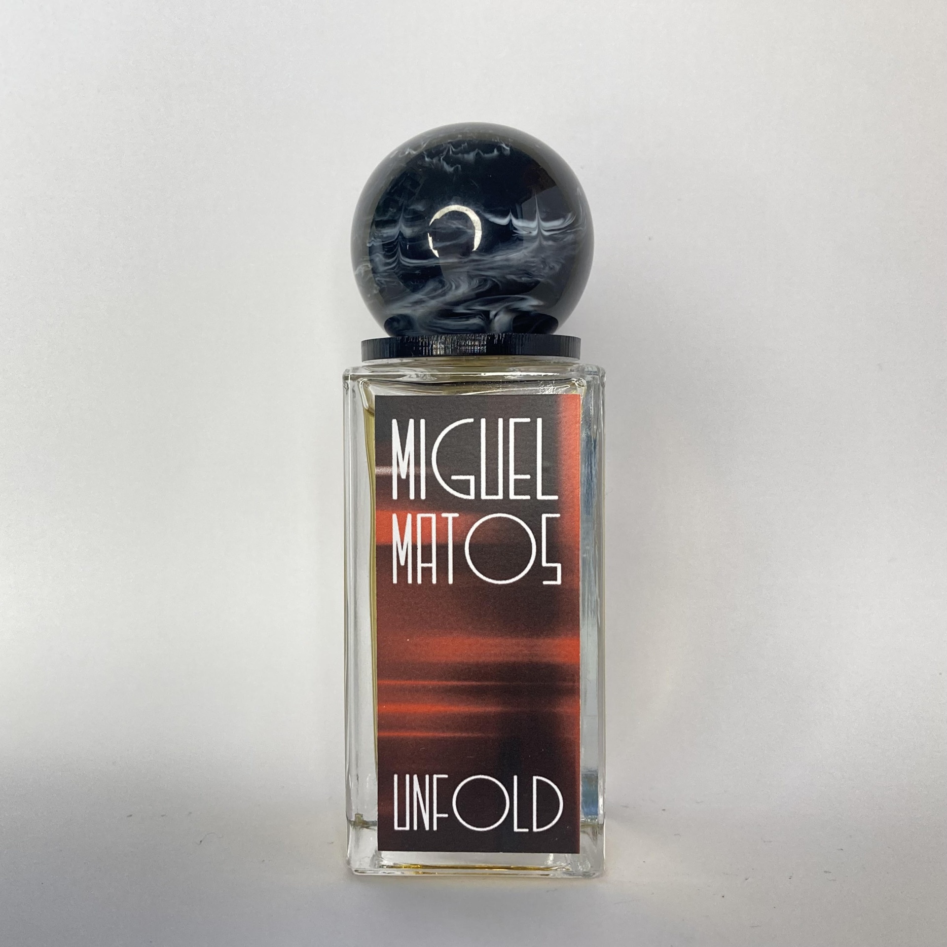 Frasco de perfume transparente com tampa redonda preta e etiqueta com texto MIGUEL MATOS UNFOLD