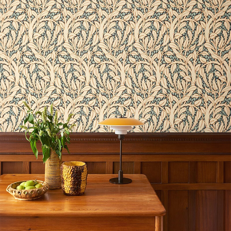 Papel de Parede Art Nouveau Fiona Wall Design | 600726 Mesa de madeira clara com plantas, vaso decorativo, tigela com maçãs e lâmpada, parede com papel de parede de folhas azuis e madeira escura.