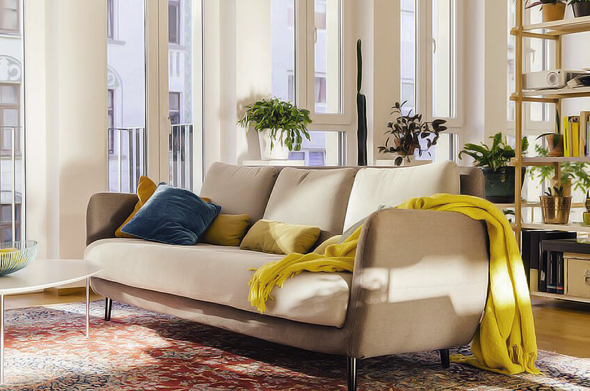 sofá bege com almofadas e manta amarela numa sala iluminada com plantas e estante