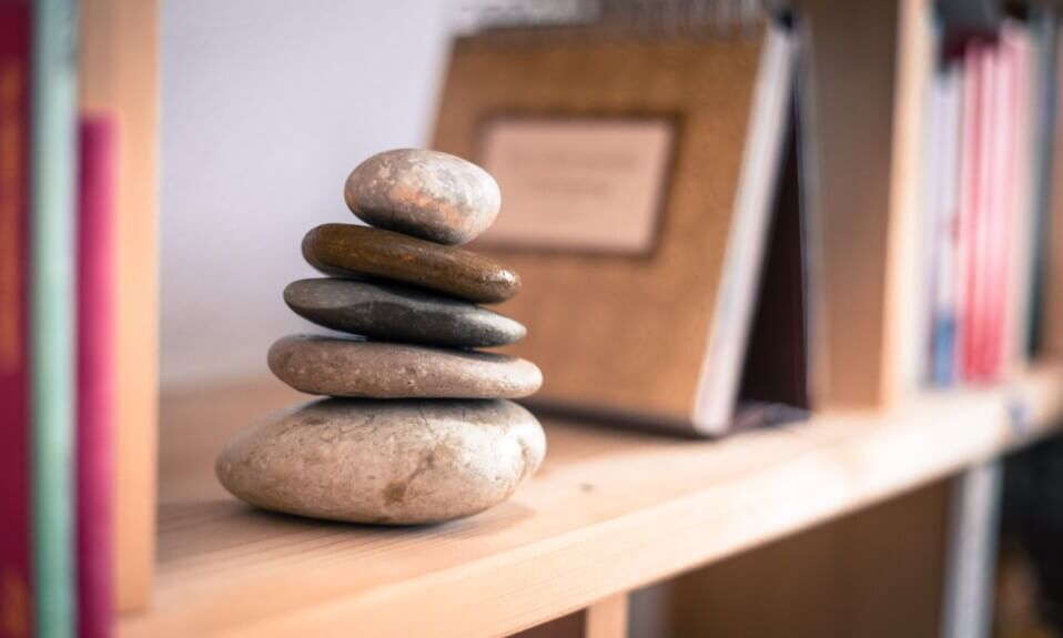 Seis pedras lisas empilhadas numa prateleira de madeira com livros ao fundo.