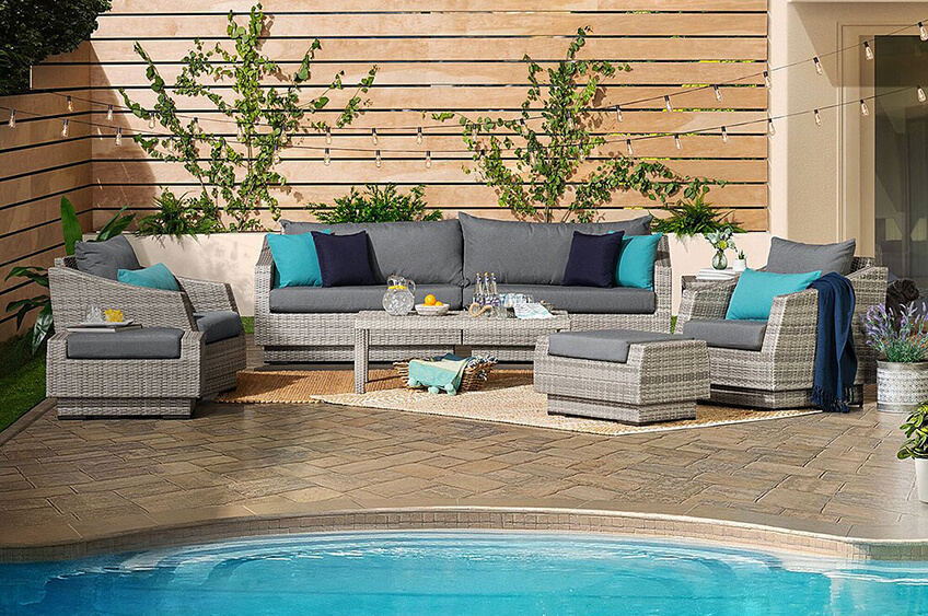 Conjunto de mobiliário de exterior em rattan sintético cinza com almofadas coloridas junto a piscina e cerca de madeira