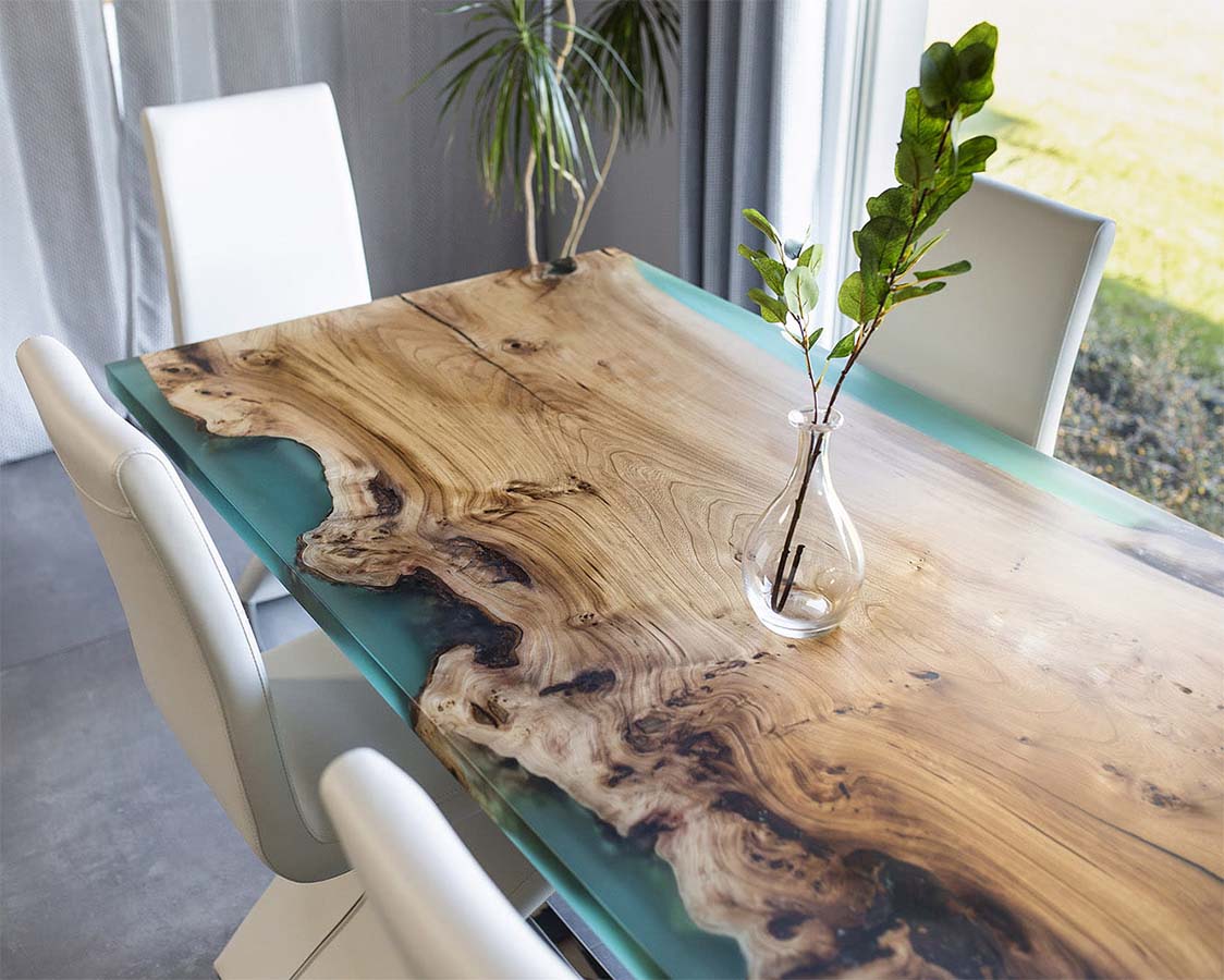 Mesa de jantar de madeira natural com resina azul e cadeiras brancas numa sala luminosa