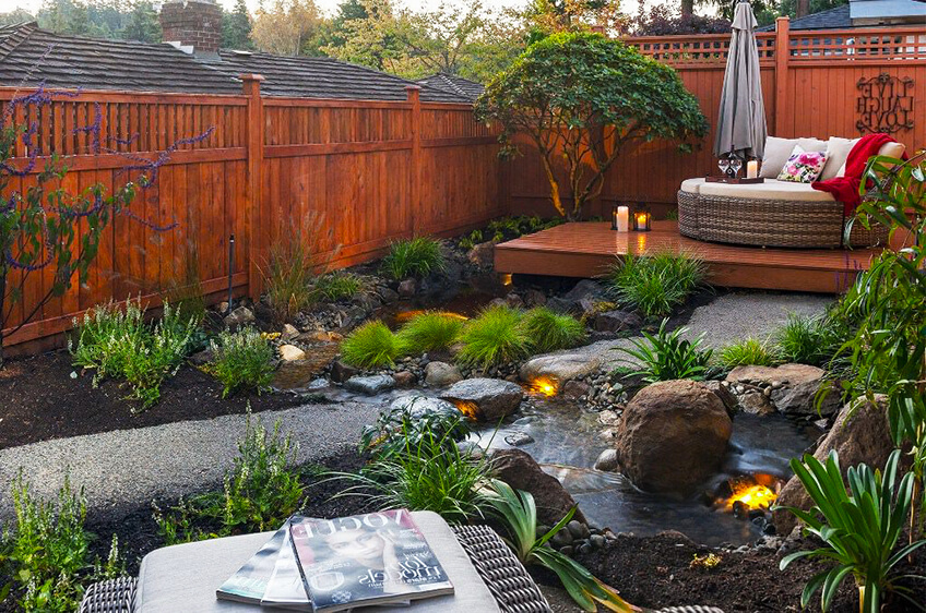 Jardim exterior decorado com cerca de madeira, plantas, riacho com pedras iluminadas, espreguiçadeira e zona de estar com banco de vime e almofadas