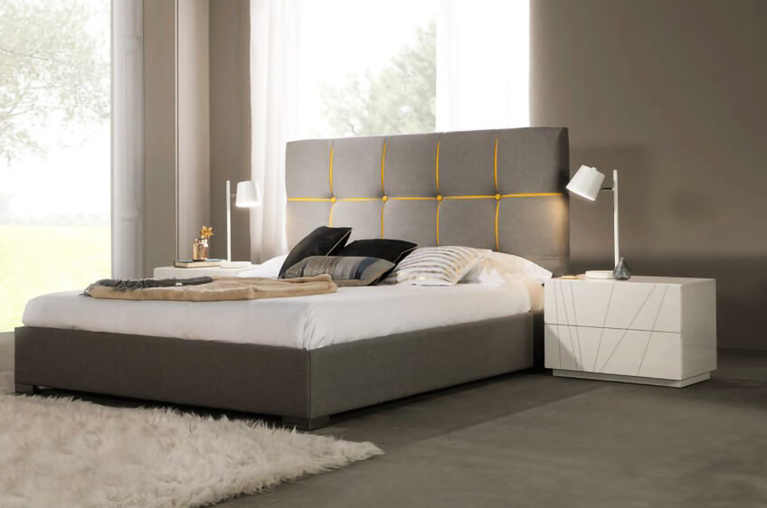 Cama moderna cinza com cabeceira detalhada em amarelo, almofadas e mesa de cabeceira branca com candeeiro.