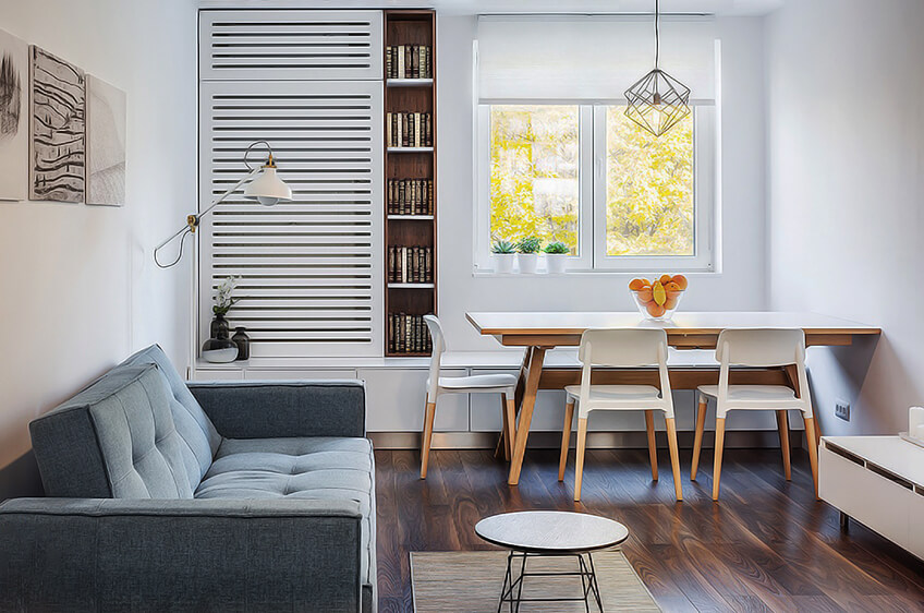Sala de estar com sofá cinza, mesa de jantar em madeira clara, cadeiras brancas e estante com livros