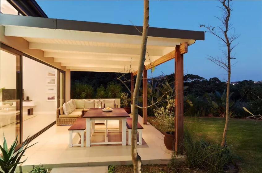 Espaço exterior com mesa de madeira rústica, bancos brancos e sofá de vime com almofadas claras