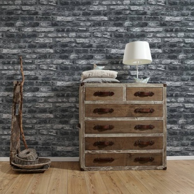 Cómoda de madeira envelhecida com lâmpada branca e almofadas, junto a aspirador antigo, parede de tijolos cinzentos.