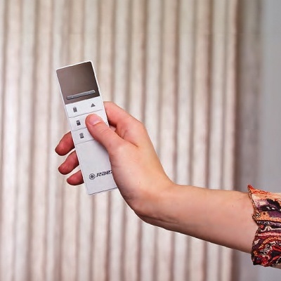 Comando remoto branco e castanho segurado por mão com manga colorida, cortinas bege ao fundo